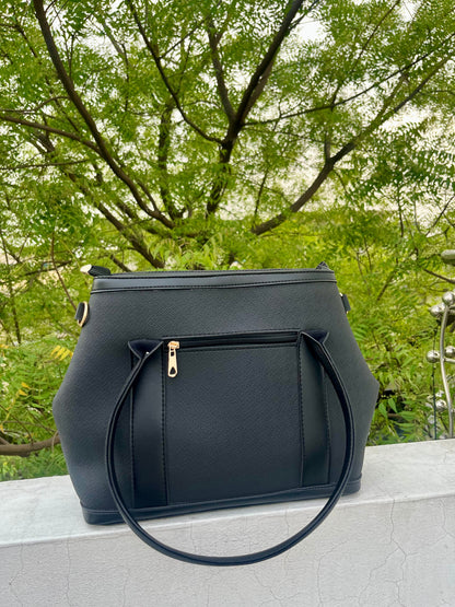 Graciza black Aero shoulder bag with flared wing design, faux leather texture, and gold hardware against green trees.