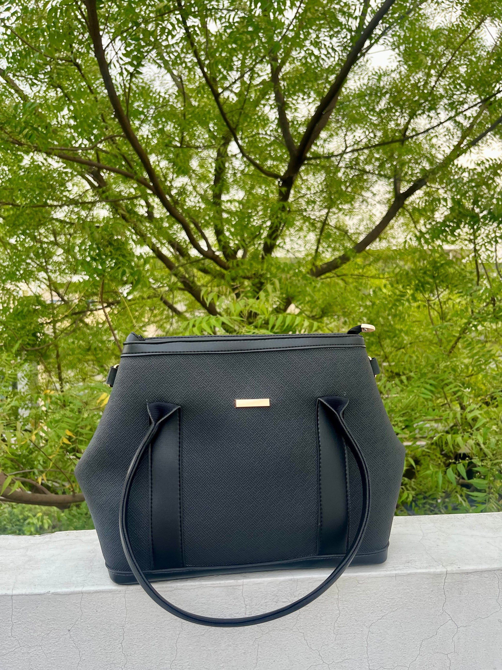 Graciza black Aero shoulder bag with flared wing design, faux leather texture, and gold bar hardware outdoors.
