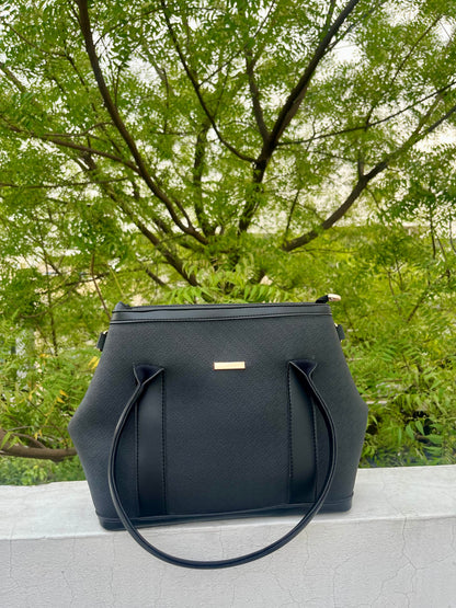 Graciza black Aero shoulder bag with flared wing design, faux leather texture, and gold bar hardware outdoors.