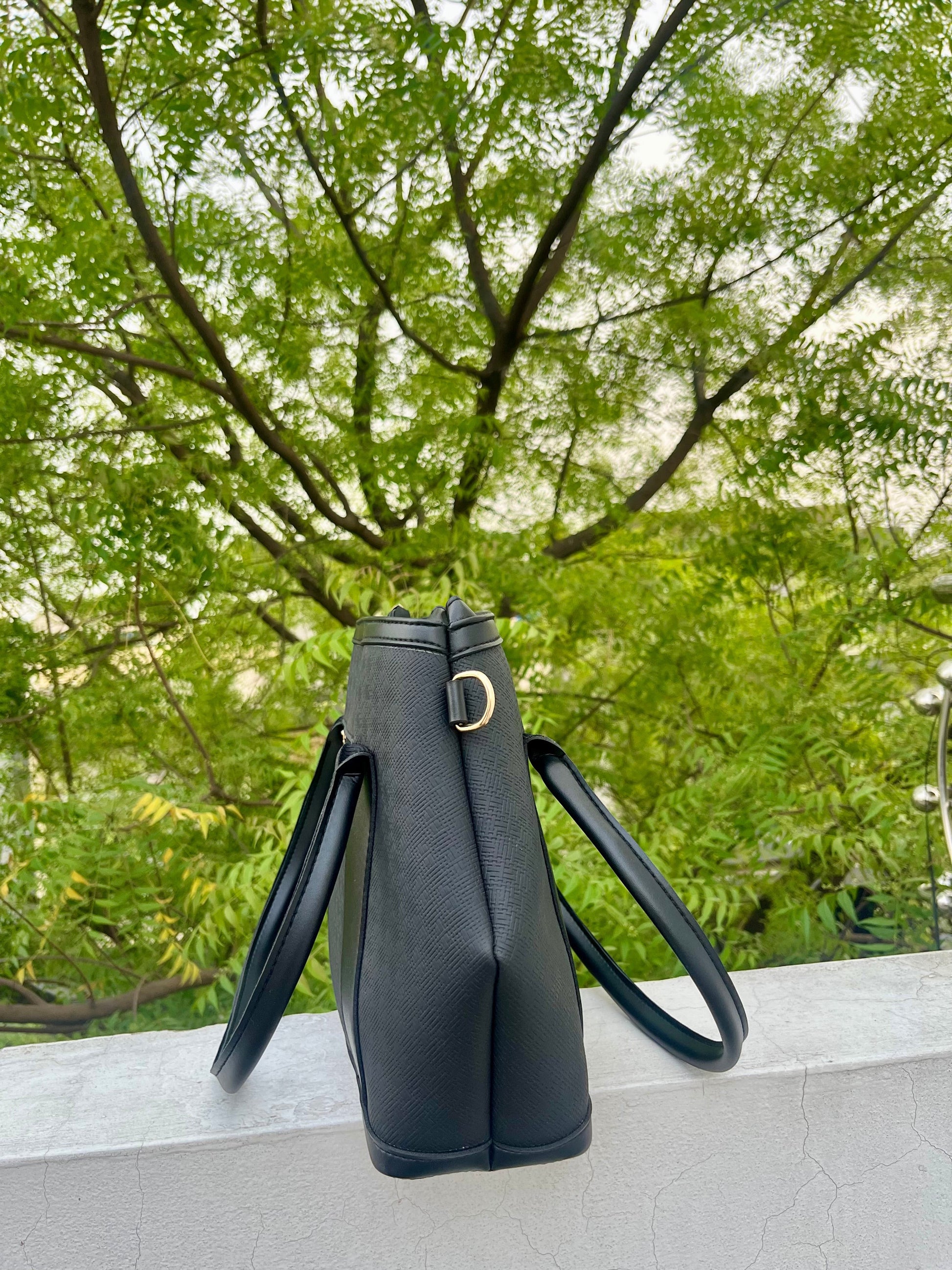 Graciza black Aero shoulder bag with flared wing design and textured faux leather finish against a green tree backdrop.
