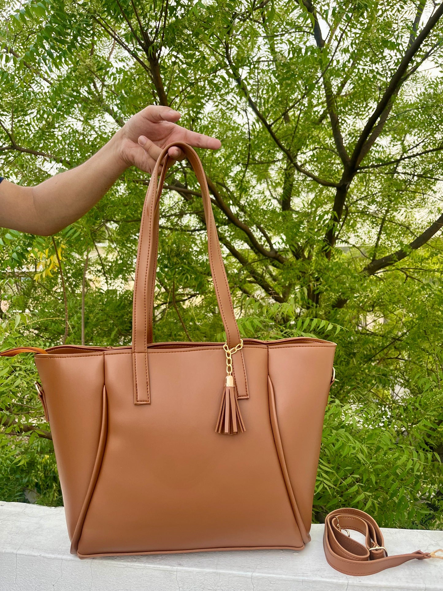 Graciza brown faux leather Aura Tote with gold hardware, decorative tassel, and removable strap against green trees.