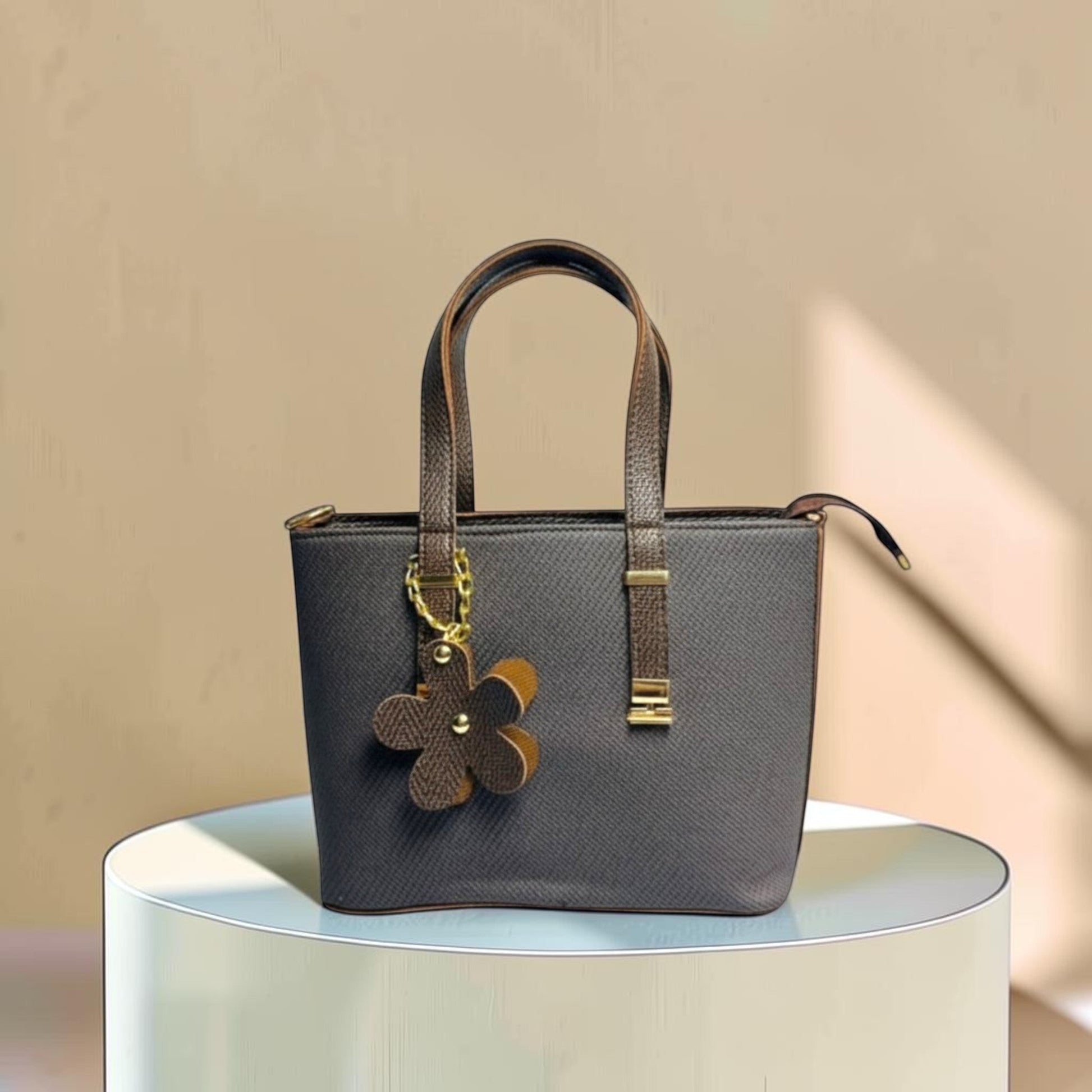 Graciza brown faux leather handbag with twin handles, zip closure, and a decorative floral charm on a display stand.