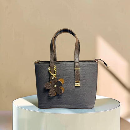 Graciza brown faux leather handbag with twin handles, zip closure, and a decorative floral charm on a display stand.