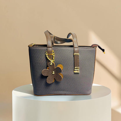 Graciza brown faux leather handbag with twin handles, zip closure, and a decorative floral charm on a pedestal.