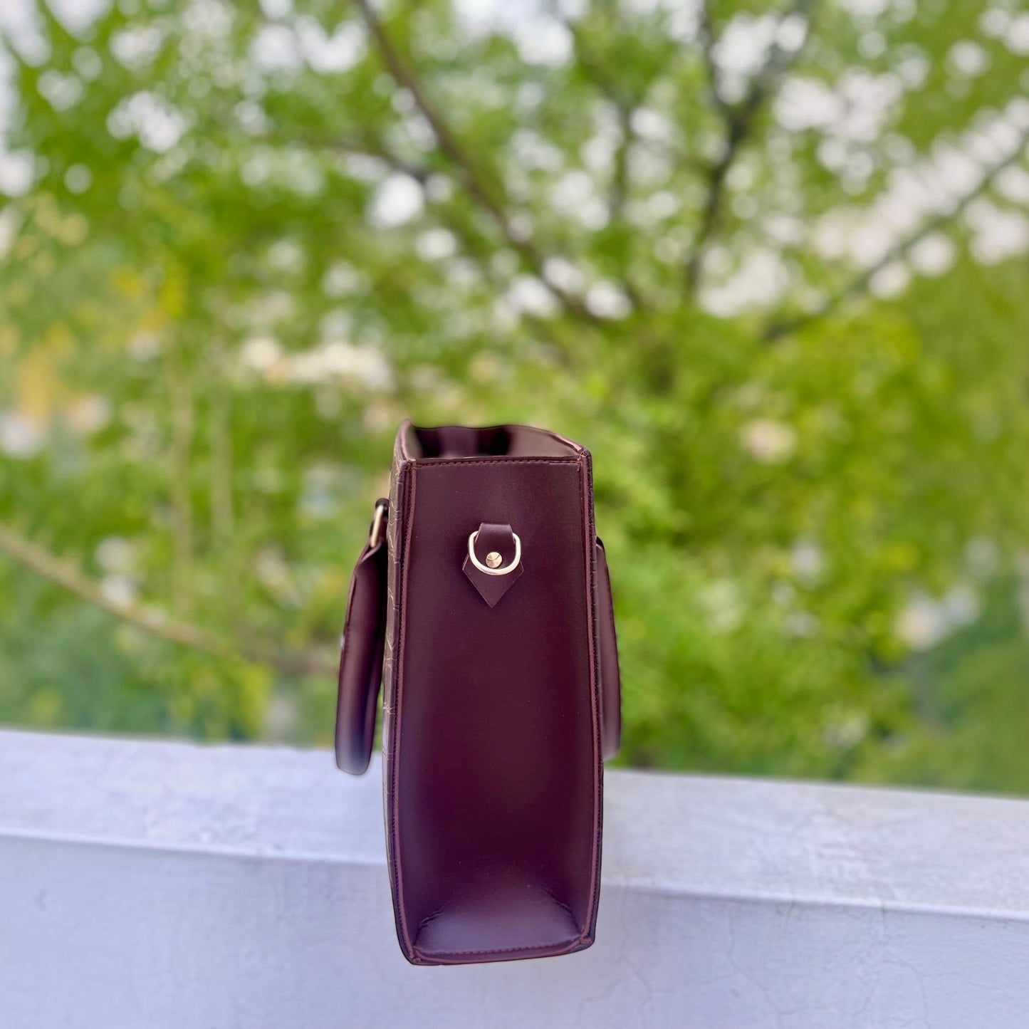 Graciza Lyra burgundy faux leather handbag with gold hardware side profile against a blurred outdoor background