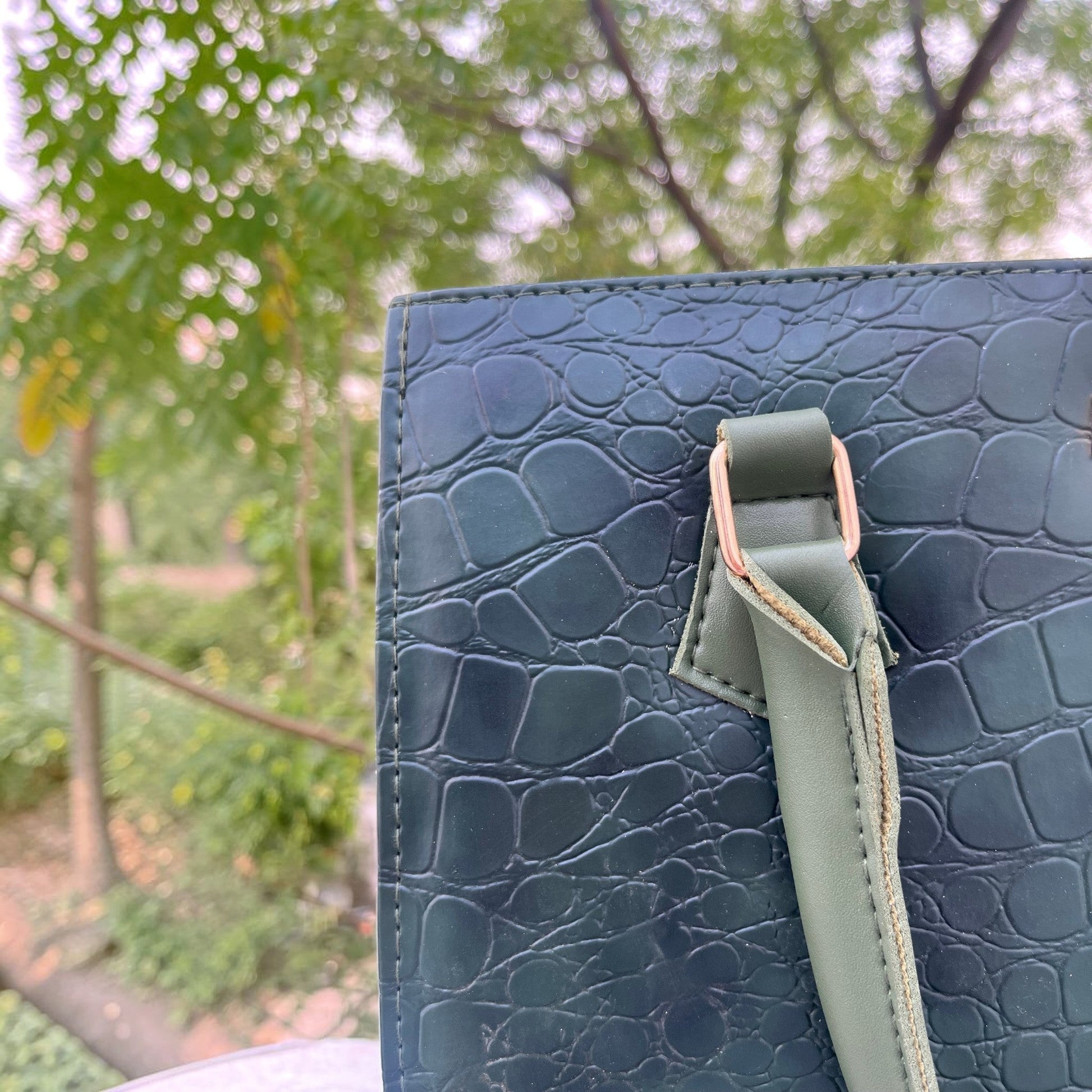 Graciza Lyra green faux leather handbag with crocodile texture and gold hardware detail against a blurred garden.